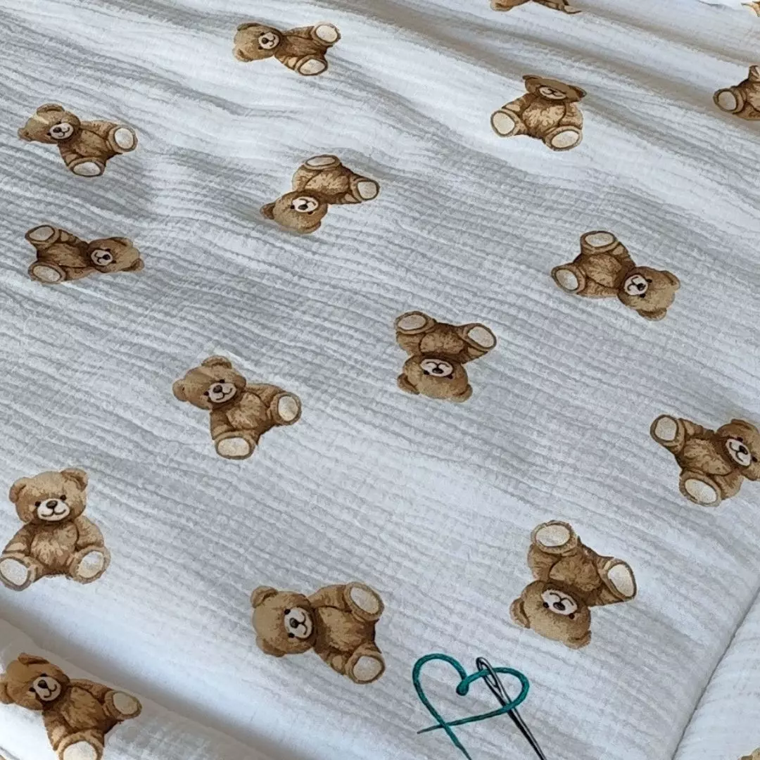 Zbliżenie na pofalowaną, kreszowaną fakturę muślinu bawełnianego Double Gauze. Śmietankowe tło z uroczym wzorem w klasyczne brązowe niedźwiadki z kolekcji Bajkowe Misie oraz widocznym u dołu subtelnym wydrukowanym logo w kształcie serca.