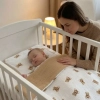 Kobieta pochylająca się nad śpiącym niemowlęciem w białym łóżeczku. Dziecko jest przykryte dwustronną pościelą muślinową we wzór w brązowe misie z widocznym ciemnobeżowym spodem.
