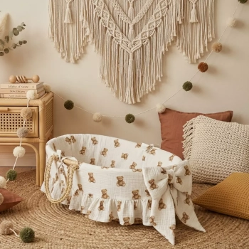 Kosz Mojżesza ubrany w muślinową wyściółkę z falbanką i dużą kokardą we wzór w klasyczne brązowe misie na śmietankowym tle. Kosz stoi na plecionym, jutowym dywanie w przytulnym pokoiku dziecięcym w stylu boho. Aranżację uzupełniają: okazała makrama ścienn