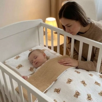 Kobieta pochylająca się nad śpiącym niemowlęciem w białym łóżeczku. Dziecko jest przykryte dwustronną pościelą muślinową we wzór w brązowe misie z widocznym ciemnobeżowym spodem.
