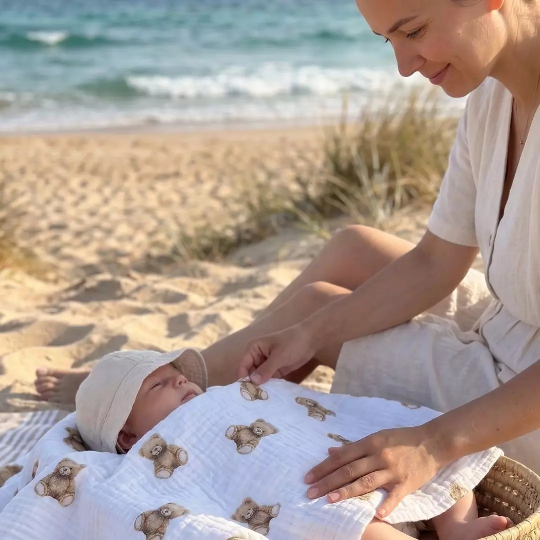 Zdjęcie lifestylowe na słonecznej plaży: uśmiechnięta mama delikatnie okrywa dziecko śmietankowym otulaczem muślinowym z kolekcji Bajkowe Misie. Kadr ukazuje naturalną, pofalowaną fakturę materiału, idealnie sprawdzającą się jako letnie, przewiewne okrycie.