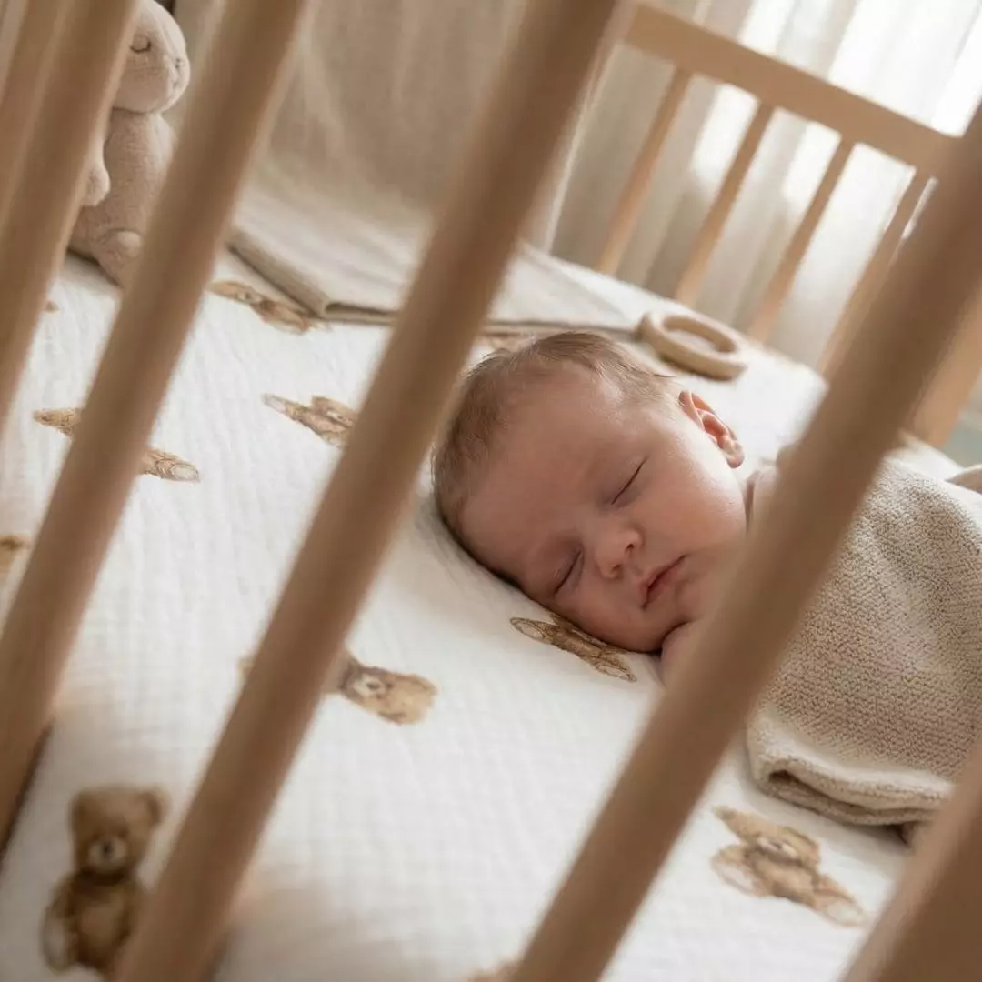Niemowlę śpiące w drewnianym łóżeczku na muślinowym prześcieradle z gumką Bajkowe Misie. Widok przez szczebelki na biały materiał w brązowe niedźwiadki.