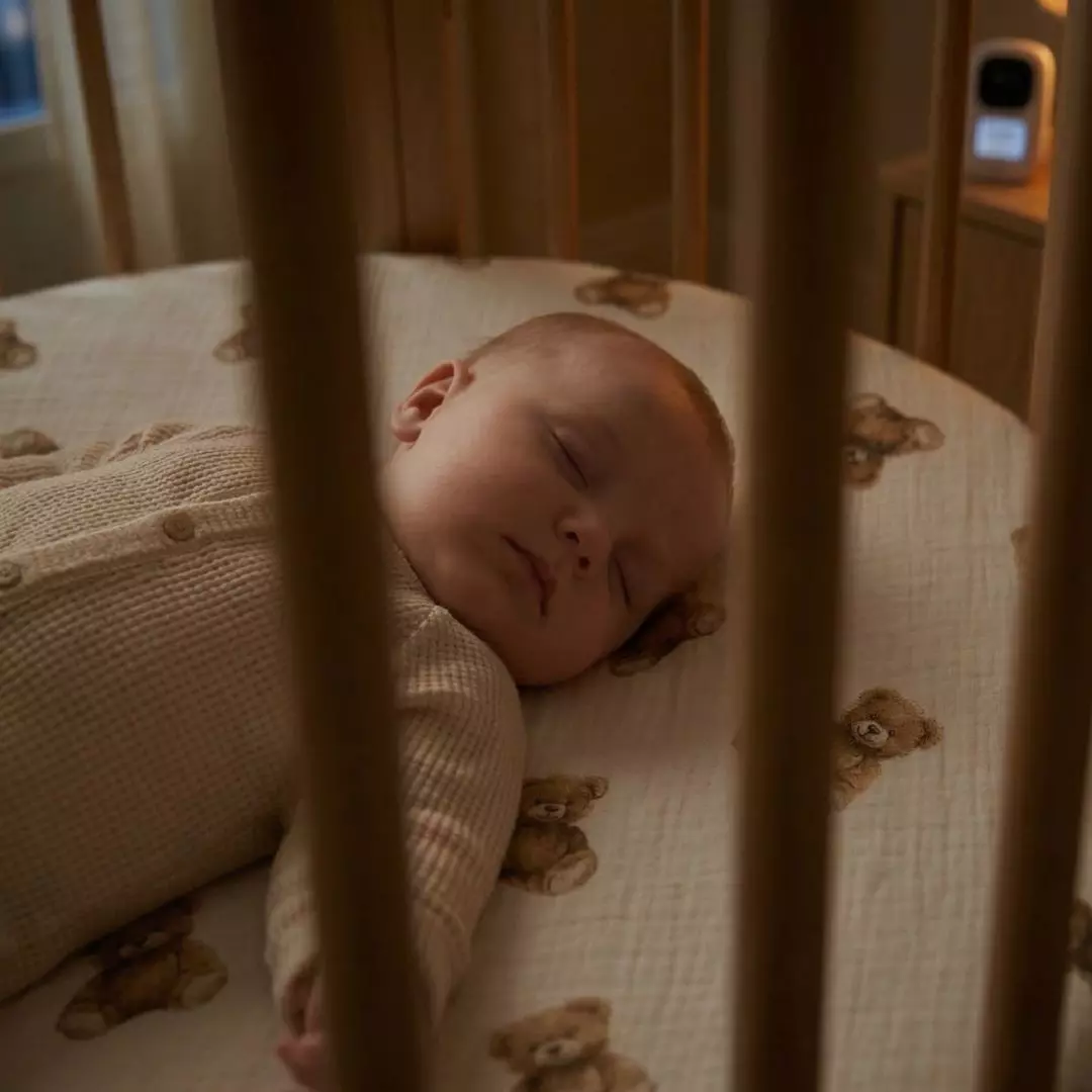 Widok przez drewniane szczebelki na śpiące niemowlę w okrągłym łóżeczku. Maluszek leży na jasnym, muślinowym prześcieradle z gumką we wzór w drobne, brązowe niedźwiadki z kolekcji Bajkowe Misie. W rozmytym, przyciemnionym tle na szafce widoczna jest elektroniczna niania.