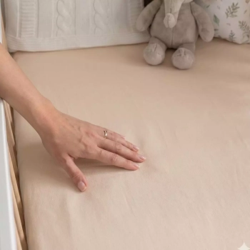 Kobieca dłoń dotykająca miękkiego, jasnobeżowego prześcieradła z gumką w łóżeczku dziecięcym. Zbliżenie na gładką strukturę materiału, w tle pluszowy słoń i dekoracyjne poduszki.