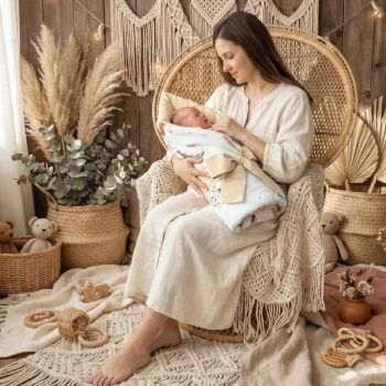 Kobieta trzymająca niemowlę w rożku, siedząca w fotelu wiklinowym w pokoju w stylu boho.