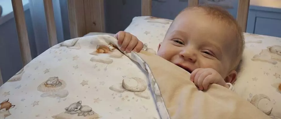 Szczęśliwe dziecko w łóżeczku pod pościelą Leśne Sny. Widoczna dwustronna pościel – wzorzysta strona w zwierzątka i gładki beżowy spód.