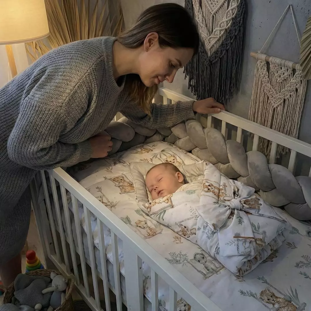 Mama z czułością patrząca na niemowlę śpiące w łóżeczku. Dziecko otulone jest w rożek z kolekcji Kołysanka z Safari, w tle welurowy ochraniacz warkocz i makramy boho