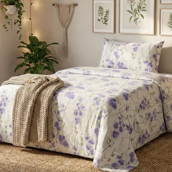 Aranżacja przytulnej sypialni: łóżko z pościelą w fioletowe akwarelowe kwiaty i zielone pnącza z kolekcji Lila Meadow. Wnętrze w naturalnym stylu boho z grubym wełnianym kocem, jutowym dywanem, roślinami doniczkowymi i makramą na ścianie.