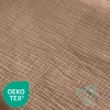 Zbliżenie na pofalowaną fakturę ciemnobeżowego muślinu bawełnianego. W lewym dolnym rogu widoczny jest zielony znak certyfikatu Oeko-Tex, a w prawym dolnym rogu turkusowe logo firmy w kształcie serduszka z igłą i nitką.