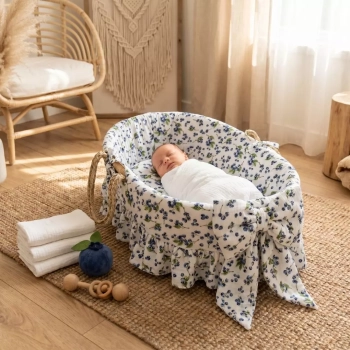 Aranżacja pokoiku w stylu boho ze śpiącym dzieckiem w koszu Mojżesza. Kosz ozdobiony jest muślinową wyściółką z falbanką i dużą kokardą oraz prześcieradłem w botaniczny wzór Leśne Jagódki.
