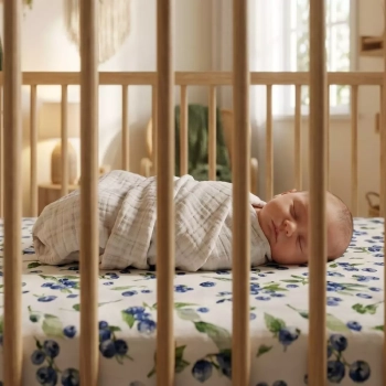 Śpiący noworodek w drewnianym łóżeczku na prześcieradle muślinowym we wzór leśne jagódki.