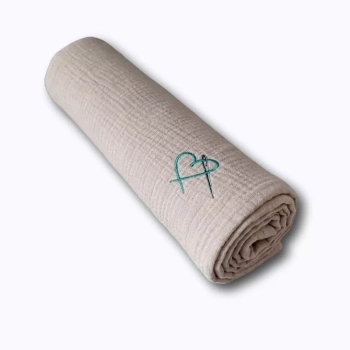 Zwinięty w rulon letni kocyk muślinowy dla niemowlaka w odcieniu jasnego beżu. Klasyczny packshot na białym tle z wyeksponowanym logo przedstawiającym serce i igłę.
