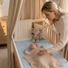 Mama w jasnym, dzierganym swetrze pochyla się z troską nad śpiącym niemowlęciem w białym łóżeczku. Łóżeczko zaścielone jest jasnoniebieskim prześcieradłem z gumką, a dziecko przykryte beżowym kocykiem. W tle widoczny jest lniany baldachim, pluszowy słonik