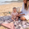 Dziewczynka w opasce siedzi na plaży na rozłożonym dwustronnym kocyku niemowlęcym. Widoczny kwiatowy wzór Retro Bloom na bawełnie i odchylony muślinowy spód w kolorze brudnego różu. Obok siedzi uśmiechnięta mama.