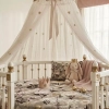 Eleganckie, okrągłe białe łóżeczko ze złotymi detalami, na którym naciągnięte jest bawełniane prześcieradło z gumką w kwiatowy wzór Retro Bloom. W łóżeczku znajduje się pluszowy króliczek w małej koronie, różowy kocyk, kwiecista poduszka oraz ozdobna, róż