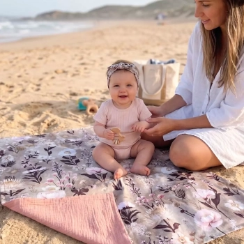 Dziewczynka w opasce siedzi na plaży na rozłożonym dwustronnym kocyku niemowlęcym. Widoczny kwiatowy wzór Retro Bloom na bawełnie i odchylony muślinowy spód w kolorze brudnego różu. Obok siedzi uśmiechnięta mama.