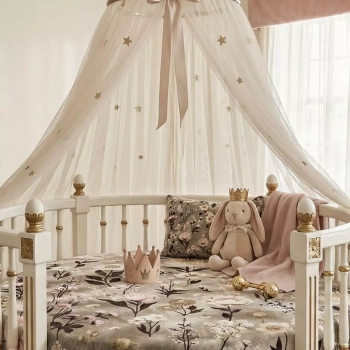 Eleganckie, okrągłe białe łóżeczko ze złotymi detalami, na którym naciągnięte jest bawełniane prześcieradło z gumką w kwiatowy wzór Retro Bloom. W łóżeczku znajduje się pluszowy króliczek w małej koronie, różowy kocyk, kwiecista poduszka oraz ozdobna, róż