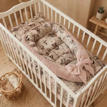 Dwustronny kokon niemowlęcy Retro Bloom leżący na kwiatowej kołderce w białym łóżeczku szczebelkowym. Wykończenie z różowego weluru z ozdobną kokardą.