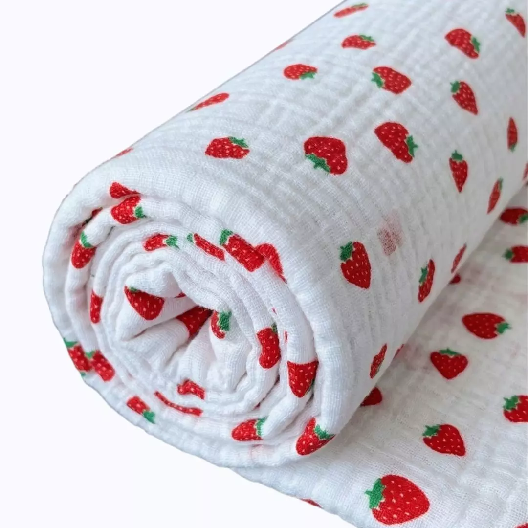 Zbliżenie na zwiniętą rolkę białej, drobnopomarszczonej tkaniny muślinowej z gęstym, powtarzalnym wzorem małych, czerwonych truskawek z zielonymi listkami. Czyste, białe tło.